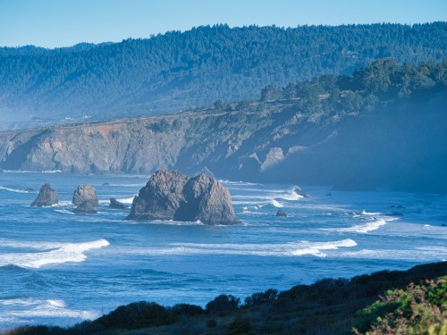 A serene coastal scene with rocky formations, gentle waves, and a misty backdrop of green hills.
