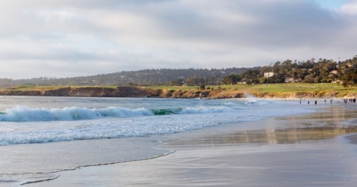 A serene beach scene with gentle waves, sandy shore, and a lush green landscape in the background under a cloudy sky.