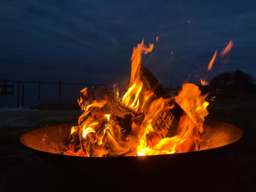 A vibrant campfire burns brightly in a metal fire pit against a darkening sky.