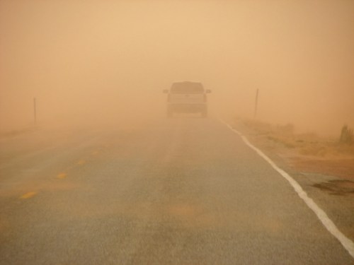 A vehicle drives down a deserted road shrouded in thick dust and fog, creating a hazy, atmospheric scene.