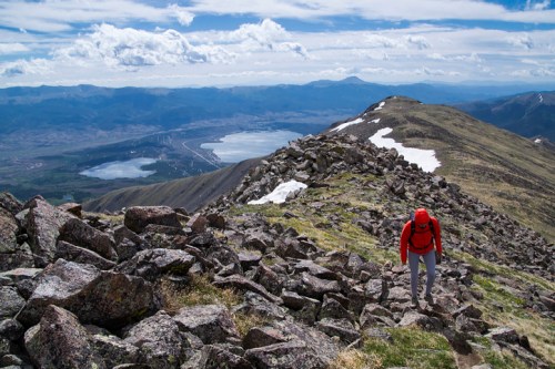 A hiker in a red jacket climbs a rocky slope with a scenic view of lakes and mountains in the background.