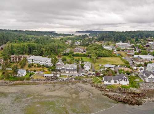 Aerial view of a coastal town with houses, greenery, and a cloudy sky over the shoreline and distant hills.