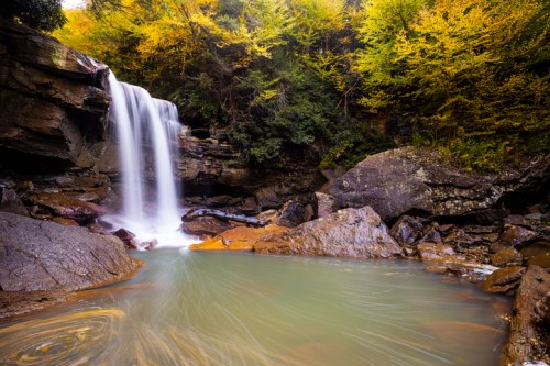A serene waterfall cascades over rocks into a calm pool, surrounded by vibrant autumn foliage.