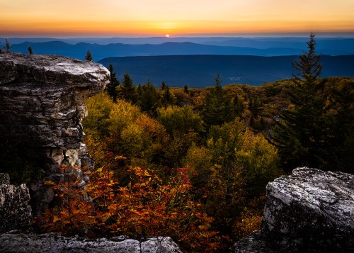 A stunning sunset over rolling mountains, framed by colorful autumn foliage and rocky outcrops in the foreground.