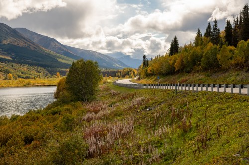 Scenic landscape featuring a winding road beside a lake, surrounded by mountains and autumn foliage under a cloudy sky.