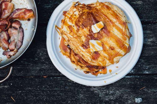 A stack of pancakes drizzled with syrup and topped with butter, alongside a plate of crispy bacon.