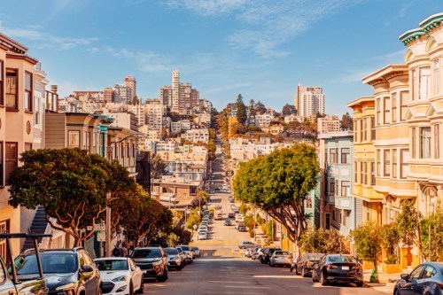 A scenic view of a hilly street lined with trees and colorful houses, leading to a city skyline under a clear blue sky.