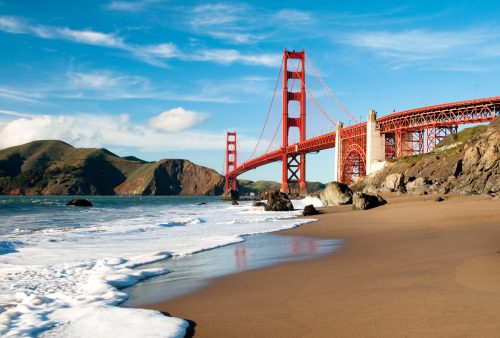Golden Gate Bridge spanning a bay, with sandy beach and waves in the foreground, under a clear blue sky.