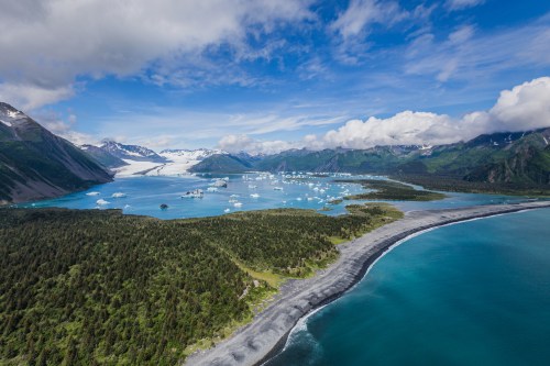 Aerial view of a stunning coastal landscape with turquoise waters, icebergs, and lush green mountains under a blue sky.