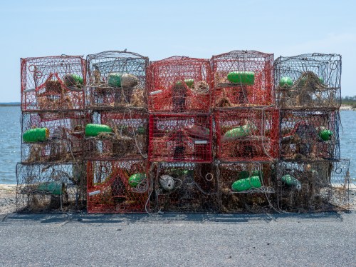 A stack of colorful lobster traps with green buoys, set against a backdrop of water and a clear sky.