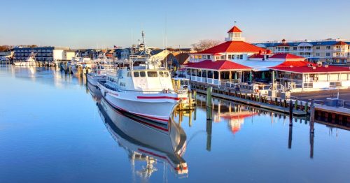 A calm marina scene with boats docked and a red-roofed building reflecting in the water under a clear blue sky.