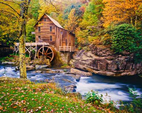 A rustic wooden mill with a water wheel beside a flowing river, surrounded by vibrant autumn foliage.