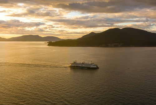 A ferry sails across calm waters at sunset, with mountains silhouetted in the background under a colorful sky.