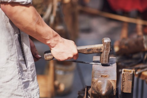 A close-up of a blacksmith's hand striking metal with a hammer on an anvil in a workshop setting.