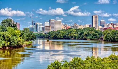 A scenic view of a city skyline reflecting on a river, surrounded by lush greenery and a blue sky with clouds.