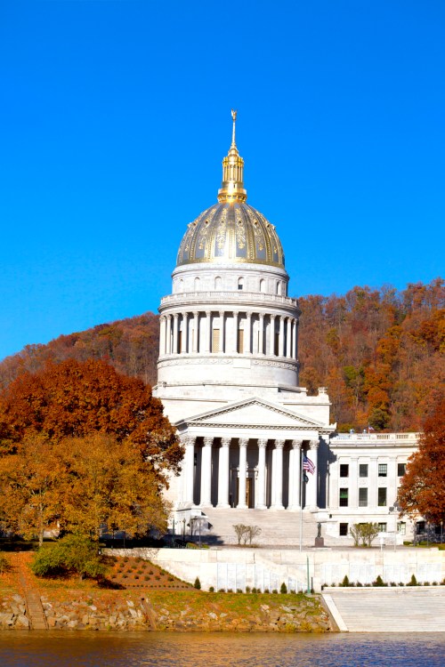 The West Virginia State Capitol building with a golden dome, surrounded by autumn foliage and a clear blue sky.