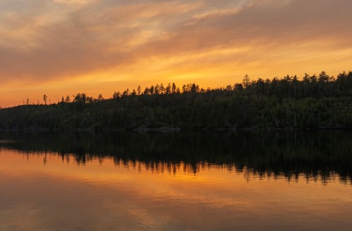 A serene sunset over a calm lake, reflecting vibrant orange and yellow hues against a forested shoreline.