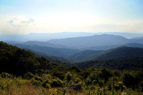 A panoramic view of rolling mountains under a clear sky, with lush greenery in the foreground and distant peaks.