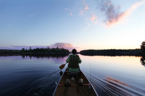 A person paddles a canoe on a calm lake at sunset, surrounded by trees and a colorful sky.