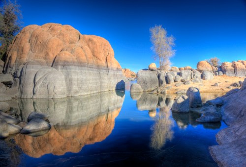 Rocky formations and a bare tree reflect in calm water under a clear blue sky.
