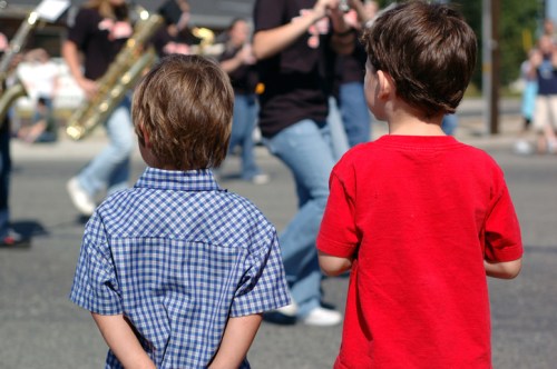 Two young boys watch a parade, one in a blue checkered shirt and the other in a red shirt, with a marching band in the background.