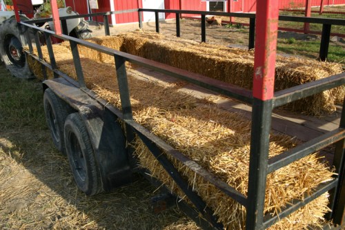 A flatbed trailer filled with bales of straw, parked on a farm with a red barn in the background.