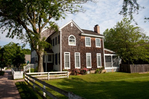 A charming two-story house with a shingled exterior, surrounded by greenery and a white picket fence.
