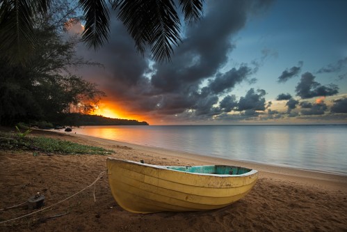 A yellow boat rests on a sandy beach at sunset, with calm waters and dramatic clouds in the background.