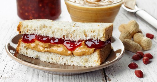 A peanut butter and jelly sandwich on a plate, with peanuts and jars of jelly and peanut butter in the background.
