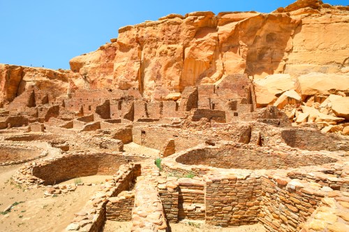 Ancient stone ruins surrounded by sandy terrain and towering cliffs under a clear blue sky.