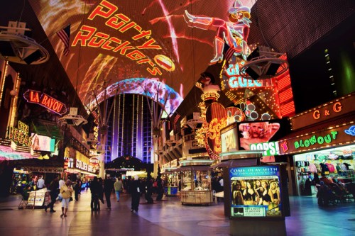 Vibrant Las Vegas street scene with neon lights, shops, and crowds under a colorful canopy.