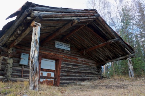 A rustic log cabin with a slanted roof, featuring a sign that reads "U.S. Post Office" and surrounded by trees.