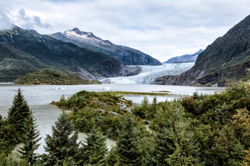 A scenic view of a glacier surrounded by mountains, with a calm lake and lush greenery in the foreground.