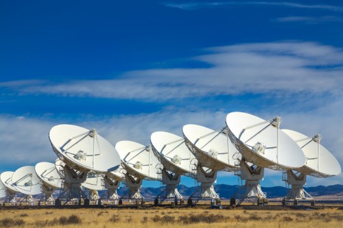 A row of large satellite dishes against a blue sky with clouds, set in a grassy landscape.