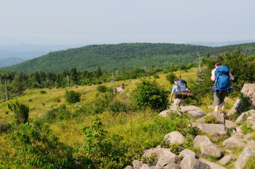 Two hikers ascend a rocky trail through a lush green landscape, with rolling hills in the background under a clear sky.