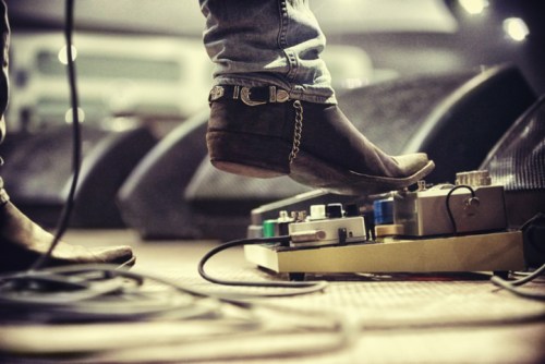 A close-up of a foot in a boot pressing a guitar pedal on stage, with cables and amplifiers in the background.