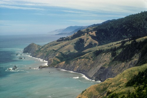 A scenic coastal view featuring rolling hills, cliffs, and the ocean under a cloudy sky.