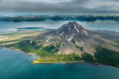 Aerial view of a snow-capped volcano surrounded by lush greenery and a calm sea, with distant mountains in the background.