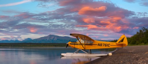 A yellow seaplane parked on a sandy shore at sunset, with mountains and colorful clouds in the background.