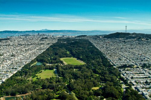 Aerial view of a city with a large park, surrounded by urban development and distant hills under a clear blue sky.