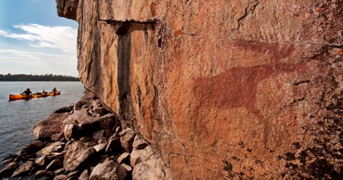 Rock face with ancient petroglyph of a moose, kayakers in the background on a calm waterway under a blue sky.