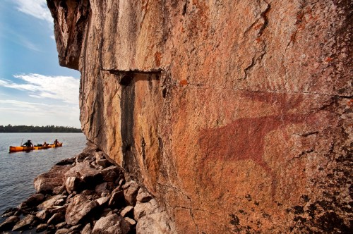 Ancient rock art depicting a moose on a cliff, with kayakers in the water nearby.