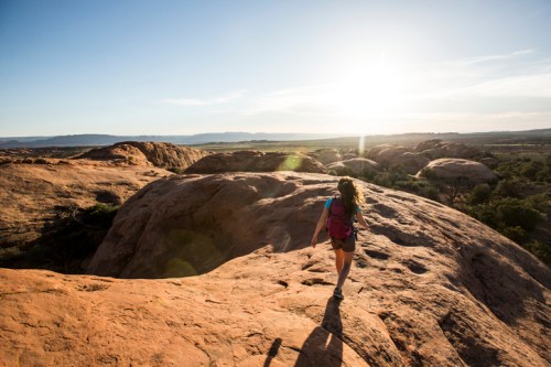 A hiker walks on rocky terrain under a bright sun, with expansive landscapes in the background.