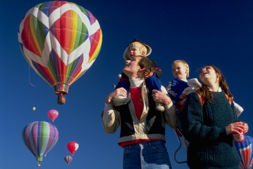 A family stands together, smiling, with colorful hot air balloons floating in a clear blue sky above them.