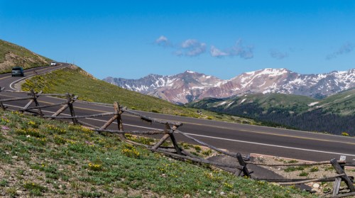A winding road through green hills, with a wooden fence and snow-capped mountains in the background under a clear blue sky.