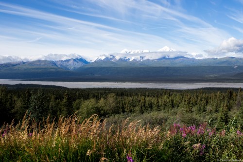A scenic view of mountains with snow-capped peaks, lush greenery, and a river under a blue sky with wispy clouds.