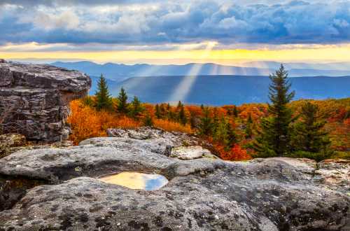 A rocky landscape with a small pool reflecting the sky, surrounded by autumn trees and distant mountains under dramatic clouds.