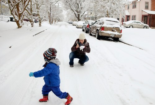 A woman crouches in the snow, taking a photo of a child running playfully in a snowy street. Snow-covered houses line the road.