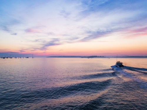 A boat glides across calm waters at sunset, with a colorful sky and distant boats in the background.