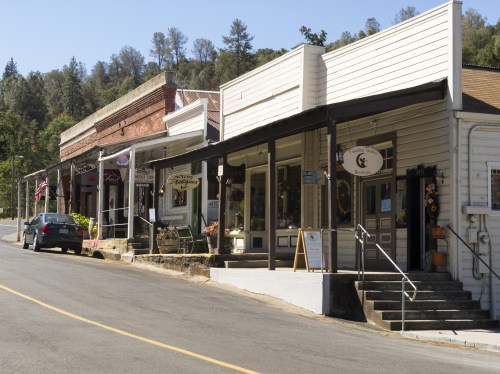 Quaint storefronts line a sunny street in a small town, with trees and hills in the background.